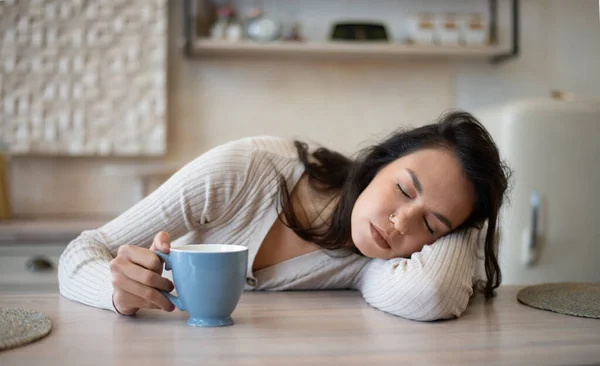 Tired woman with coffee cup sleeping on table in modern light kitchen interior, lying on table with closed eyes. Overwork at home, facial expression in early morning and breakfast alone
