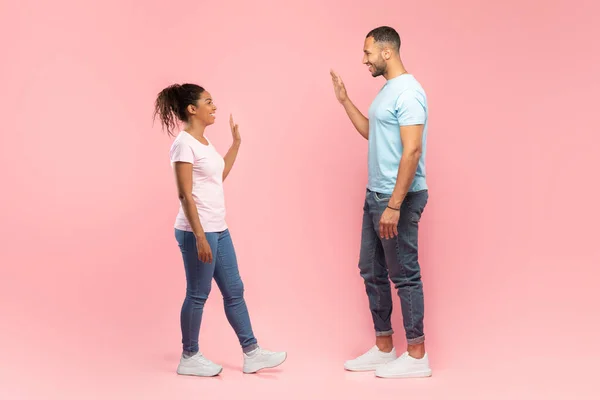 Excited black man and woman meeting each other and waving hands, posing over pink studio background, side view. Friendship, relationship and communication concept. Full length shot