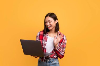 Çevrimiçi video görüşmesi. Mutlu Asyalı kadın sarı arka planda web kamerasına el sallıyor ve dizüstü bilgisayarını kullanıyor. Arkadaşlarıyla ya da ailesiyle konuşan neşeli bir kadın.