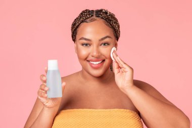 Mutlu Afro-Amerikan kadın elinde pamuk ped ve yüz losyonu ile şişe tutuyordu, pembe stüdyo arka planında duruyordu. Güzel siyah eoman, kendine bakmanın keyfini çıkarıyor.