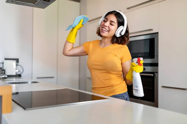 Cheerful Young Arab Woman Having Fun While Doing Cleaning In Kitchen, Millennial Middle Eastern Female Wearing Wireless Headphones Listening Favorite Music And Dancing While Tidying Home, Free Space