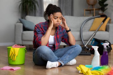 Çökmüş binlerce yıllık Afrika kökenli Amerikalı kadın oturma odasında kirli ve dağınıklıktan muzdarip temizlik malzemeleriyle yerde oturuyor. Ev işlerinden, sorunlardan, evde çalışmaktan kaynaklanan stres.