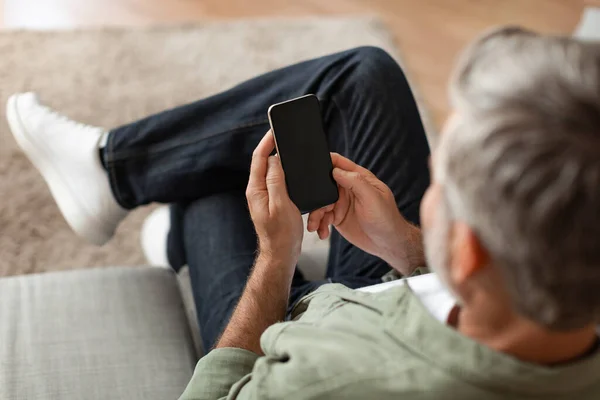 チャットのための新しいアプリを使用して空の画面でスマートフォンに入力する高齢者の白人男性は 室内のソファの上に座っています 提供のための近代的な技術 自宅でウェブサイトの推奨 — ストック写真