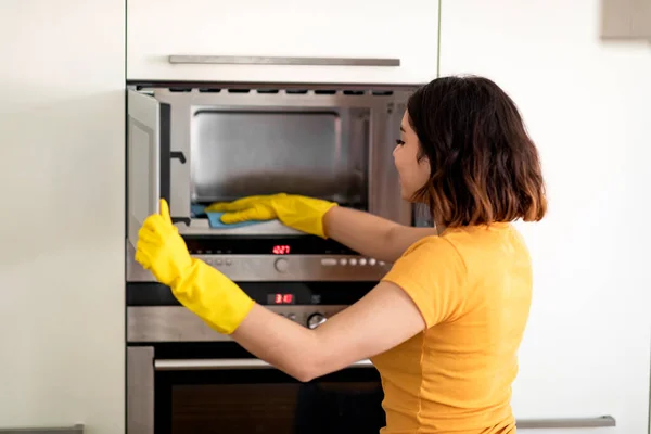 Happy Young Arab Woman In Rubber Gloves Wiping Microwave With Rag While Cleaning In Kitchen, Smiling Middle Eastern Housewife Tidying Home, Enjoying Making Domestic Chores, Closeup Shot