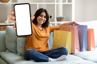 Cep telefonu reklamı. Gülümseyen Orta Doğulu Kadın Boş Akıllı Telefon Gösterisi ve Parlak Alışveriş Çantaları Evdeki koltukta otururken, Mutlu Arap Kadın Çevrimiçi Alım Uygulamalarını Öneriyor, Mockup