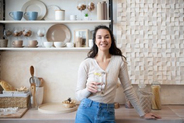 Sağlıklı hidrasyon konsepti. Serinlemek için su içen mutlu genç bayan, modern mutfağın içinde duruyor. Susuz kalma, zayıflama ve kilo verme konsepti için beslenme