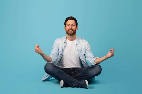 Happy calm young european bearded guy in casual meditates with closed eyes, sits on floor in lotus position, isolated on blue background, studio. Man enjoy peace and silence, lifestyle and health care