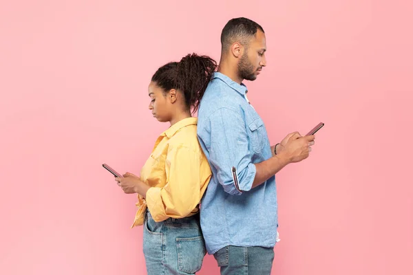 ソーシャルネットワーク中毒 アフリカ系アメリカ人のカップルがスマートフォンを使って立ち上がり 互いを無視して ピンクのスタジオの背景 サイドビュー — ストック写真