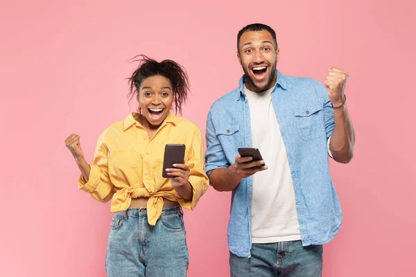 素晴らしいニュースだ アフリカ系アメリカ人のカップルがスマートフォンを持ち 成功を祝い ピンクの背景に手を上げ 興奮して拳を握りしめました — ストック写真