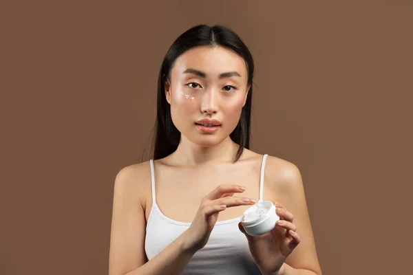 Skincare cosmetics concept. Korean lady trying new moisturising product, holding open jar with under eye cream, enjoying selfcare, and making beauty routine, posing over brown background
