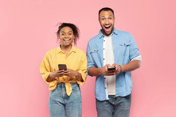 モバイルオファー 喜びに満ちた黒のカップルがスマートフォンを持ち 興奮してカメラを見ている姿 興奮した男と女がピンクの背景にポーズをとって口を開ける — ストック写真