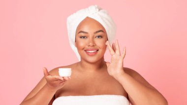Güzel Afro-Amerikan kadın cildine yüz kremi sürüyor, elinde kavanozla kameraya gülümsüyor, pembe stüdyo arka planı, panorama