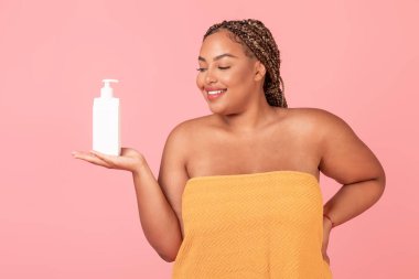 Havluya sarılmış, elinde vücut losyonuyla şişeyle duran, yeni bir cilt bakım ürünü öneren, pembe arka planda poz veren, stüdyo çekimi yapan iri yarı, güzel bir kadın. Model