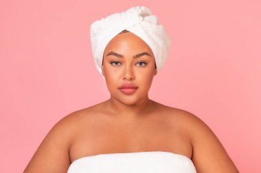Havlu giymiş, pembe stüdyo arka planında duran Afro-Amerikan kadın portresi. Kusursuz derisi olan, kameraya bakan, iri yapılı, güzel bir kadın.