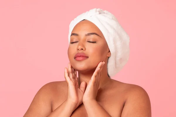 Portrait of body positive black woman with smooth skin touching her face while posing over pink studio background. Pretty plus size lady wrapped in bath towel enjoying beauty care