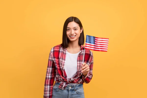 Education abroad, studying in the US, emigration concept. Happy asian female student holding american flag, posing over yellow studio background, copy space
