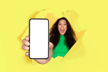 Aşırı mutlu genç kadın boş ekranlı modern akıllı telefonu gösteriyor sarı kağıt duvardaki yırtık delikten, mobil uygulama ya da web sitesi reklamı için model, kolaj