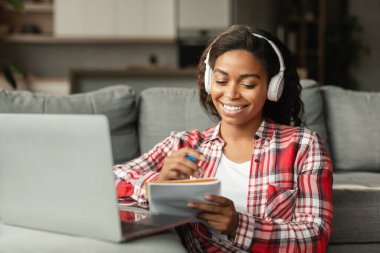 Kablosuz kulaklık takan, binyıllık siyahi bayan öğrenci oturma odasındaki bilgisayarla evde ders çalışıyor. Bilgi, modern teknolojiyle uzaktan eğitim, online ders, evde yeni normal.