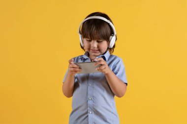Çocuklar ve online oyunlar. Kulaklık takan, akıllı telefondan video oyunu oynayan, turuncu arka planda, serbest alan kullanan küçük çocuğun stüdyo portresi.