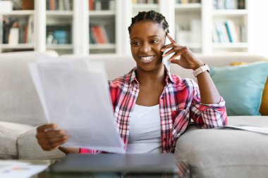 Kadınlar iş başında. Milenyum girişimcisi neşeli, başarılı, evden çalışan, oturma odasında oturan, telefon görüşmeleri yapan, belgelere bakan ve gülümseyen güzel siyah kadın.