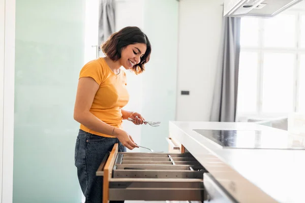Smiling Middle Eastern Female Tidying Up Cutlery In Kitchen Drawer, Side View Shot Of Happy Young Arab Housewife Assembling Forks While Doing Cleaning At Home, Making Domestic Chores, Copy Space