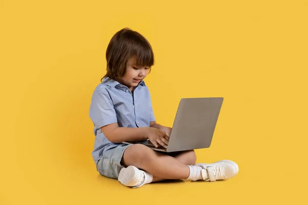Little tech master. Adorable little boy typing on laptop computer, playing developing game, sitting on floor, orange studio background