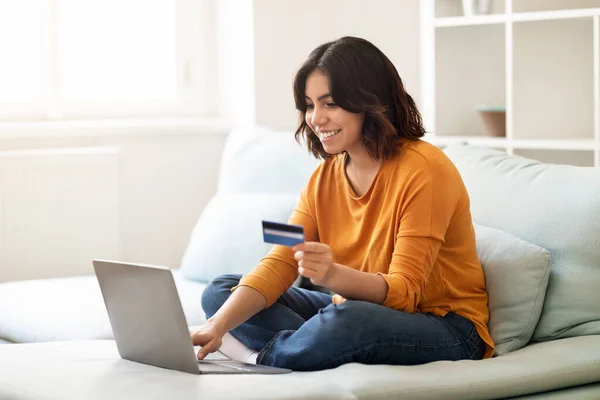 Smiling Young Arab Woman Making Online Payments With Laptop And Credit Card While Sitting On Couch At Home, Happy Millennial Middle Eastern Female Enjoying Purchasing From Internet, Copy Space