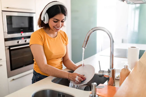 台所で料理を洗濯し 無線電話で音楽を聞きます 洗剤でハッピー中東女性クリーニングプレート 家事の雑用を楽しみます スペースをコピー — ストック写真
