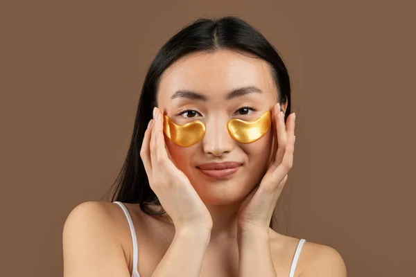 Happy korean woman applying eye patches, enjoying procedures on brown background, studio shot. Skin care, natural beauty, spa treatment concept