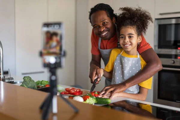 キッチンでカメラで料理をする黒人の父と娘 フードブログでビデオを録画し 健康的なサラダを一緒に準備しながら三脚でスマートフォンを使用してアフリカ系アメリカ人の家族を笑顔 — ストック写真