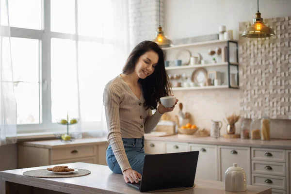 ラップトップとお茶を飲んで幸せな白人女性は キッチンのインテリアでテーブルの上に座って インターネットサーフィン 無料のスペース 朝食を取り 家から遠隔で働いている女性 — ストック写真