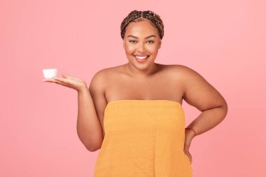 Güzellik bakımı. Çekici siyah vücut pozitifliği olan kadın yüz kremi ile kavanozu tutuyor ve kameraya gülümsüyor, havluya sarılmış duruyor, yeni kozmetik ürünleri tanıtıyor, pembe arka plan.