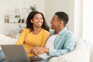 Gülümseyen bin yıllık Afro-Amerikan erkeği kadınlara bakar çevrimiçi arama için dizüstü bilgisayar kullanır, açık oturma odasında video izler. İnternette sörf, harika haberler, aşk, romantik ve evdeki insanların duyguları.