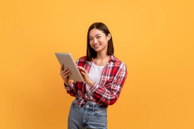 Dijital tablet kullanan ve kameraya gülümseyen mutlu Japon kadın sarı stüdyo arka planında dururken internete göz atıyor. Modern teknolojiden hoşlanan bayan