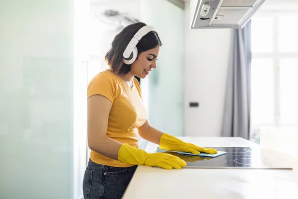 Smiling Middle Eastern Housewife Washing Table In Kitchen With Rag, Happy Young Arab Woman Wearing Wireless Headphones Doing Cleaning At Home And Enjoying Favorite Music, Side View With Copy Space