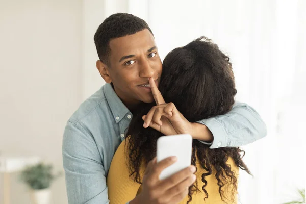 陽気千年アフリカ系アメリカ人の夫抱擁妻は沈黙のサインを表示し リビングルームのインテリア フリースペースでスマートフォンを保持します 秘密裏のジェスチャー オンライン驚き 贈り物のためのアプリ 自宅での関係 — ストック写真
