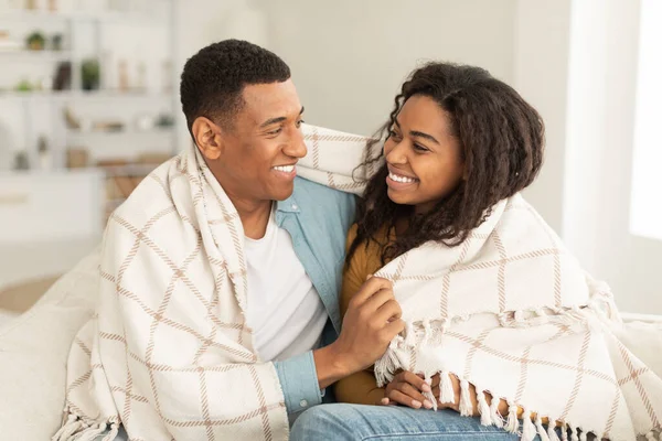 Smiling millennial african american male and female wrapped in plaid, hugging, enjoy tender moment in free time in light living room interior. Love, romance and relationships, warm and comfort at home