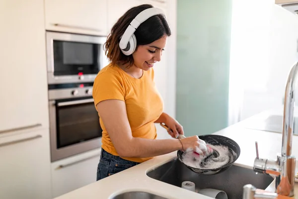 キッチンで料理をしたり ワイヤレスで音楽を聴いたりしながらパンを洗う幸せなアラブ人女性 洗剤でスポンジを使って中東の若い女性を微笑ませ 家事の雑用を楽しむ — ストック写真