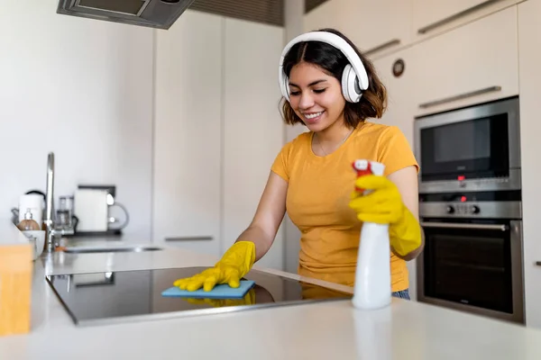 Happy Young Arab Lady Wearing Headphones Doing Cleaning In Kitchen At Home, Smiling Middle Eastern Woman Washing Induction Cooktop Surface With Rag And Listening Favorite Music, Free Space
