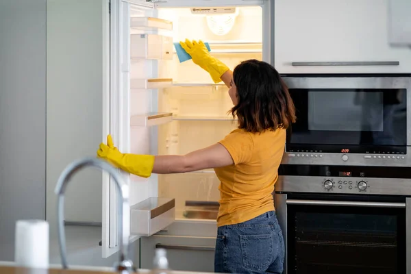 Young Housewife Wiping Shelves In Empty Fridge While Doing Cleaning In Kitchen, Smiling Female Wearing Rubber Gloves Using Rag While Tidying Home, Enjoying Making Domestic Chores, Free Space