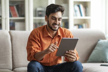 Pozitif havalı, yakışıklı, orta doğulu genç adam oturma odasında kanepede oturuyor, dijital tablet kullanıyor ve gülümsüyor, arkadaşlarıyla sohbet ediyor, web sörfü yapıyor, güzel bir uygulama kullanıyor, uzayı kopyalıyor.