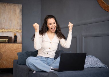 Harika haberlerim var. Başarıyı dizüstü bilgisayarla kutlayan neşeli bir bayan, evdeki koltukta oturuyor, boş yer var. Heyecanlı beyaz kadın yumruklarını kaldırıyor ve heyecanla bağırıyor.