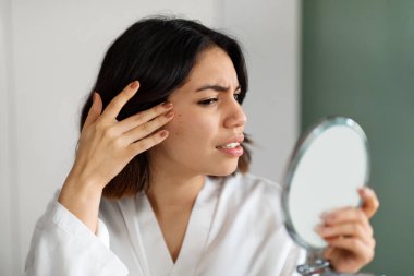 Beyaz ipek bornozlu üzgün bir orta doğulu kadın akne hastası, Arap bir kadın el aynasında cildini kontrol ediyor, yanağındaki kırmızı sivilcelere dokunuyor, kopyalama alanı. Deri bozuklukları kavramı