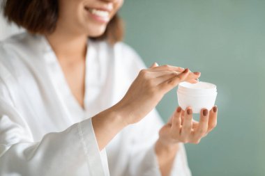 Beyaz ipek bornozlu esmer genç bir kadın evde yüz kremi kullanıyor. Yeni organik güzellik ürünlerini test ediyor. Plastik bir kavanoz tutuyor.