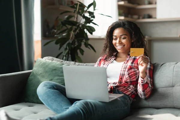 Online shopping, big sale. Smiling millennial black lady shopaholic use computer and credit card to check banking account, sit on sofa in interior living room. Cashback, finance and lifestyle at home