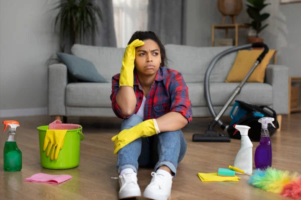 Unhappy tired millennial black lady sit on floor with cleaning supplies, suffer from mess and dirt in living room interior. Stress from household chores, wait for cleaning service, many work at home