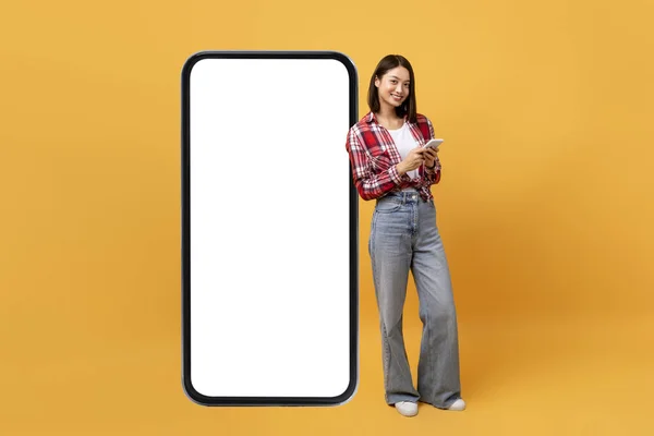 Excited korean lady using smartphone while standing near giant smartphone with blank screen on yellow studio background, full body length. Mock up for website or app, copy space for advert
