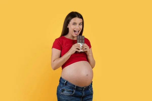 黄色の背景に隔離されながら 朗らかな若い妊婦さんがチョコレートバーを噛み カメラを見ている姿をポートレートしながら 妊娠中のお菓子を食べる幸せそうな女性 — ストック写真