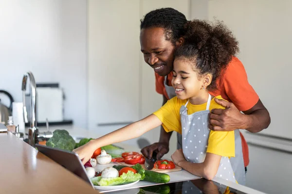 キッチンでお父さんと料理をしながらデジタルタブレットでレシピをチェックかわいい黒の女の子 家庭で昼食のために食事を作りながら 幸せなアフリカ系アメリカ人の父と娘は成分のリストを見て — ストック写真