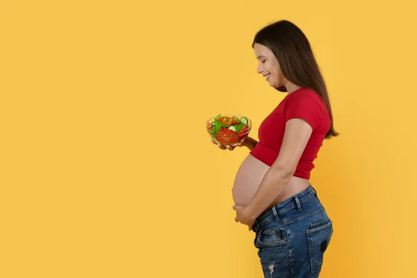 Pregnancy Nutrition. Beautiful Pregnant Woman Holding Bowl With Vegetable Salad And Caressing Belly While Standing Isolated On Yellow Background, Happy Expectant Lady Enjoying Healthy Food, Side View